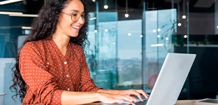 Mulher parda, com cabelo solto, usando óculos e mexendo no notebook.